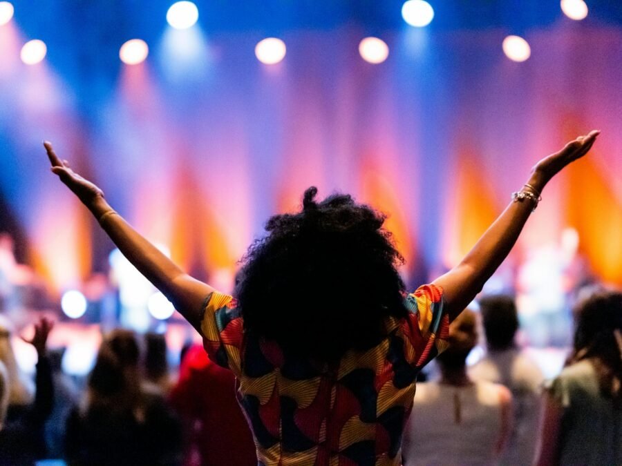 Back view of anonymous ethnic female viewer enjoying performance with arms apart against people and stage with shiny spotlights